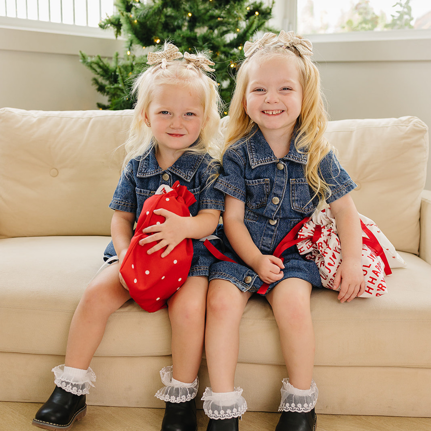 Two young girls in matching outfits sitting on a couch with Christmas decorations in the background.