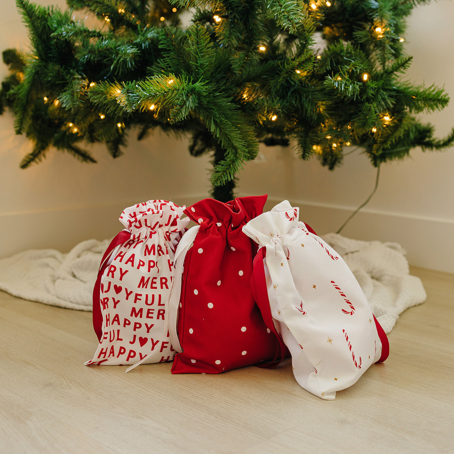 Three gift bags under a decorated Christmas tree on a wooden floor.