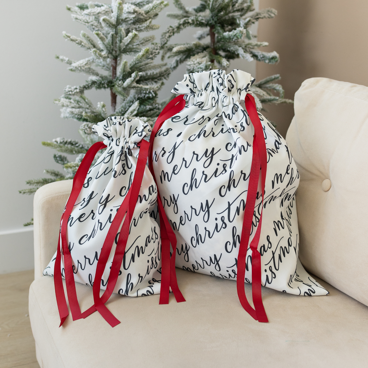 Two white fabric gift bags with 'Merry Christmas' text in black calligraphy, each adorned with a red ribbon.