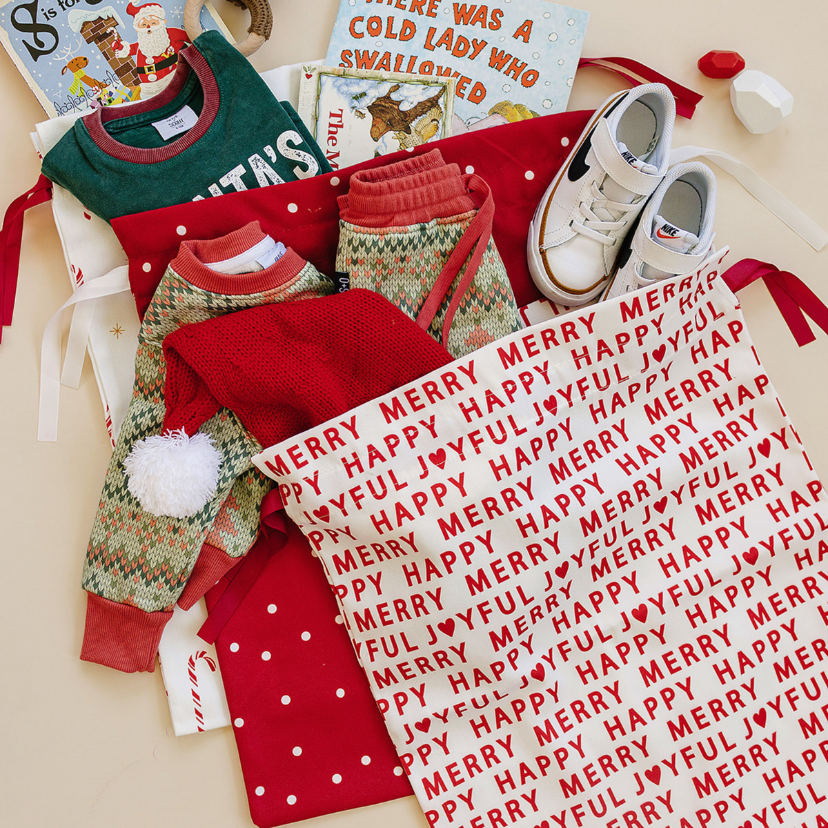 Collection of Christmas-themed items including clothing, shoes, books, and reusable fabric Christmas gift bags on a beige surface.
