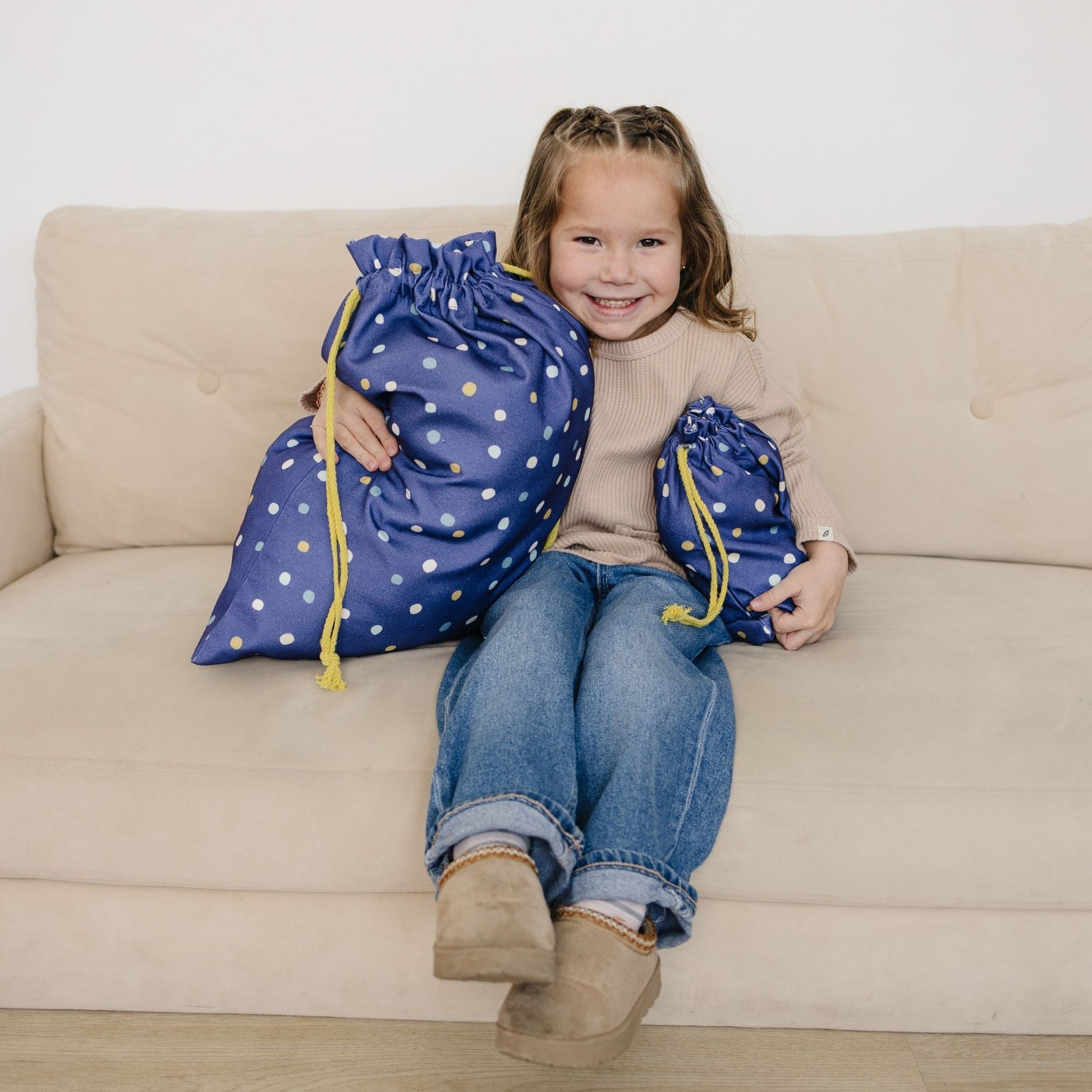 Child sitting on a couch holding two large blue drawstring bags with white polka dots.
