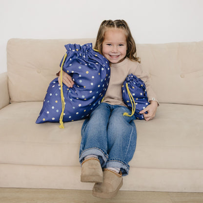 Child sitting on a couch holding two large blue drawstring bags with white polka dots.