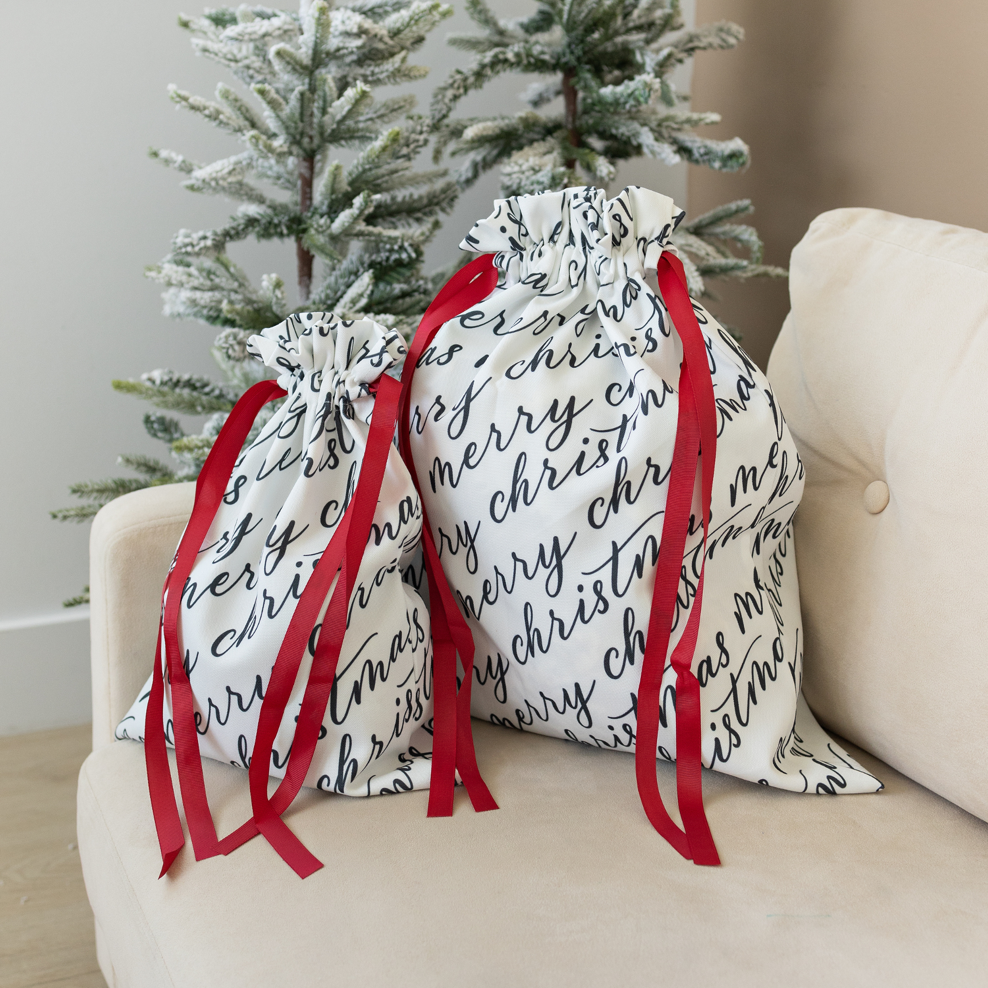 Two white fabric gift bags with 'Merry Christmas' text in black calligraphy, each adorned with a red ribbon.