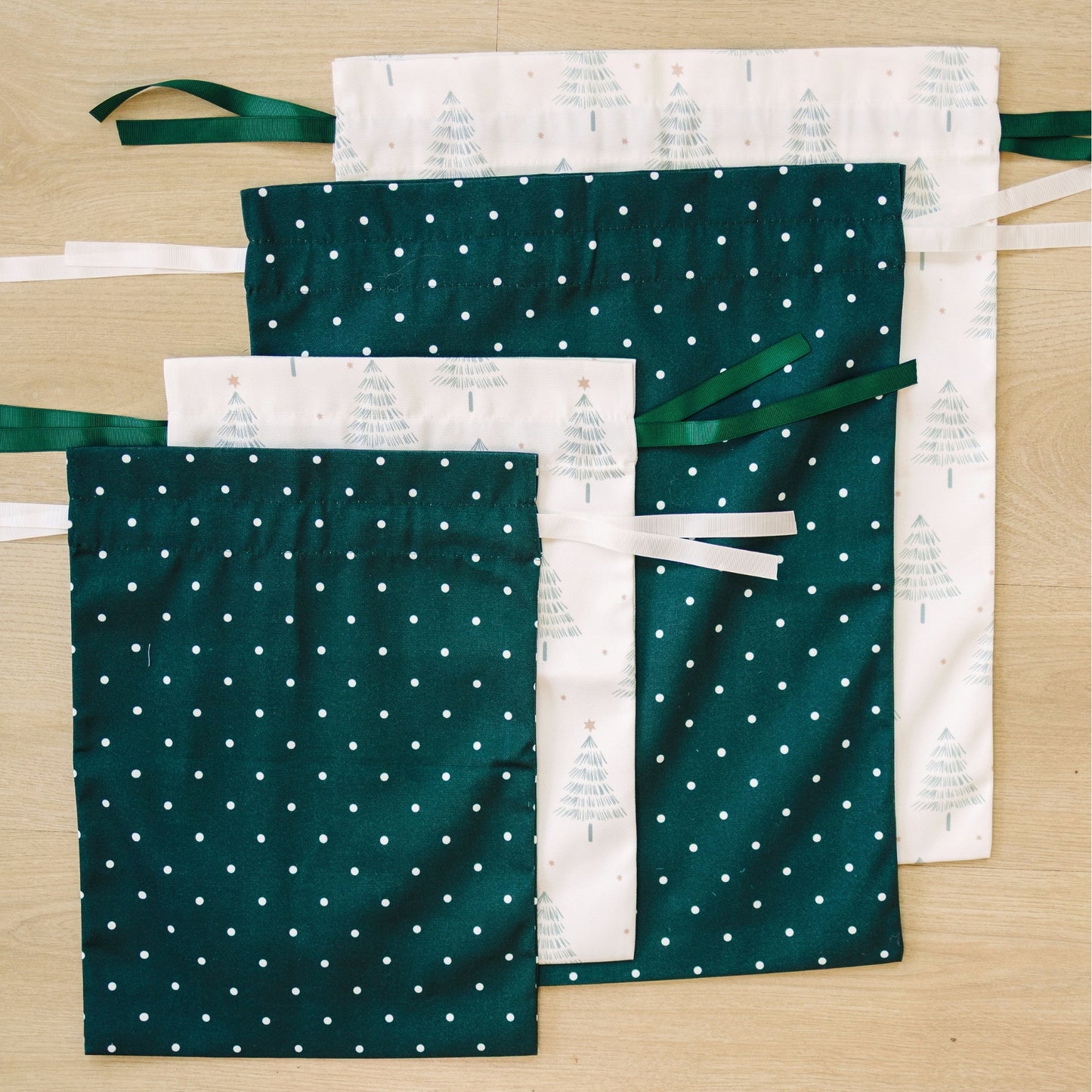 Four fabric drawstring pouches arranged on a light wood surface. Two pouches are dark green with small white polka dots, and two is white with a green tree pattern.