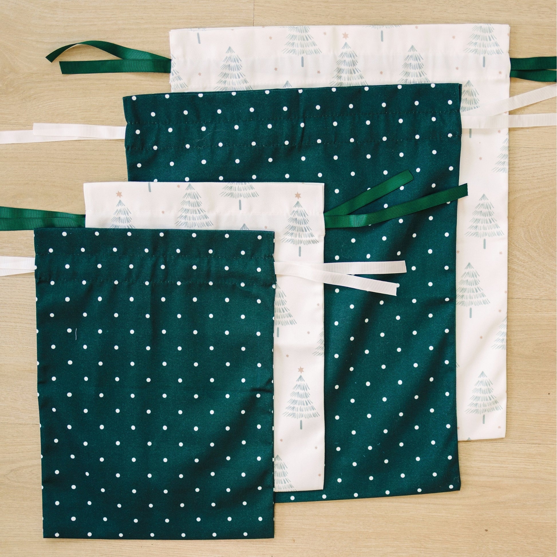 Four fabric drawstring pouches arranged on a light wood surface. Two pouches are dark green with small white polka dots, and two is white with a green tree pattern.