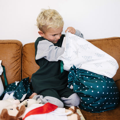 A young blonde child sits on a brown couch, opening a white patterned fabric gift bag and a dark green drawstring bag with white polka dots. 