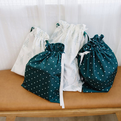 Four fabric drawstring pouches arranged on a tan bench in front of a white curtain. Two pouches are dark teal with white polka dots, and two are white with a pale tree print.