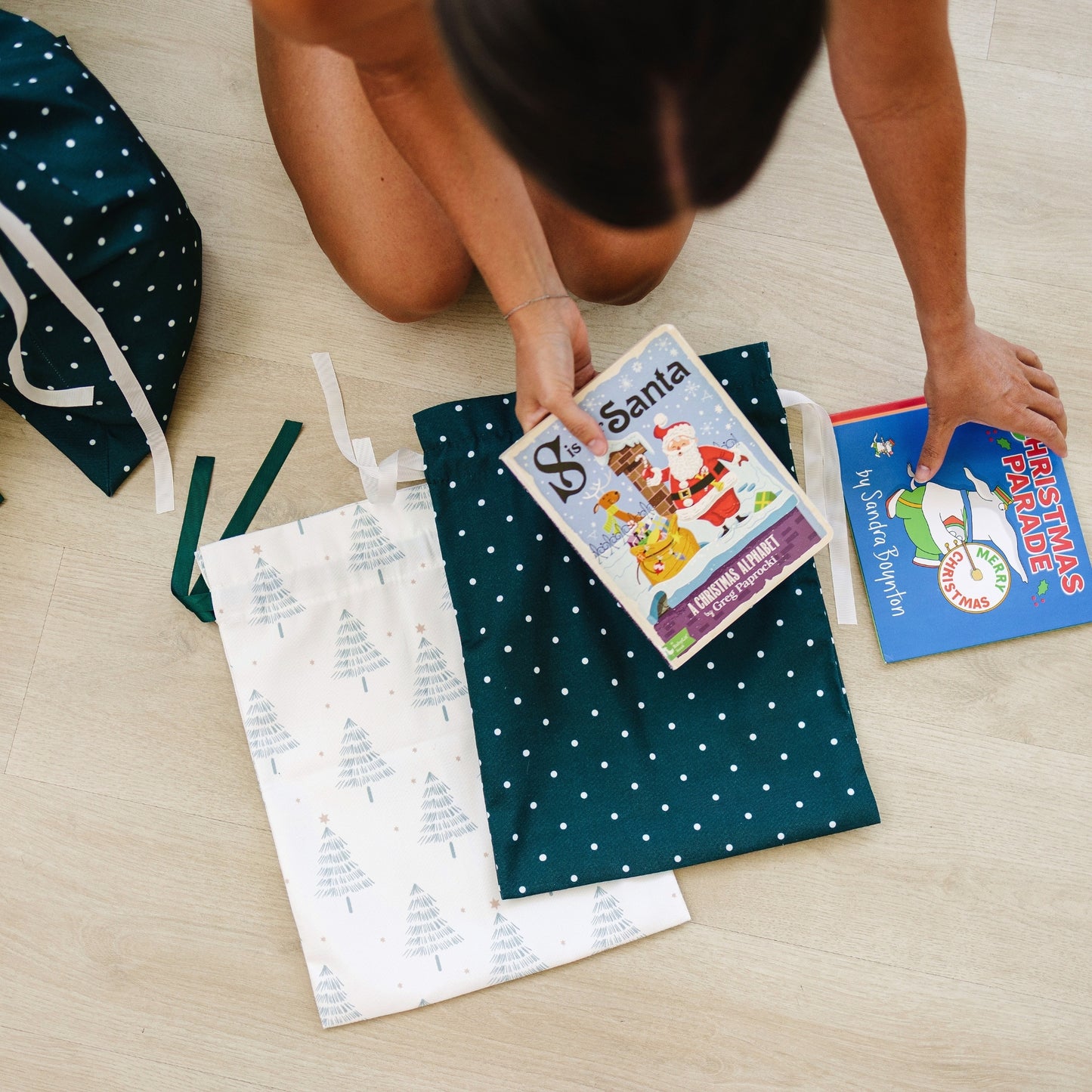 A person kneels on a light wood floor arranging Christmas-themed fabric gift bags and books. The bags include one dark green with white polka dots and one white with a Christmas tree print.
