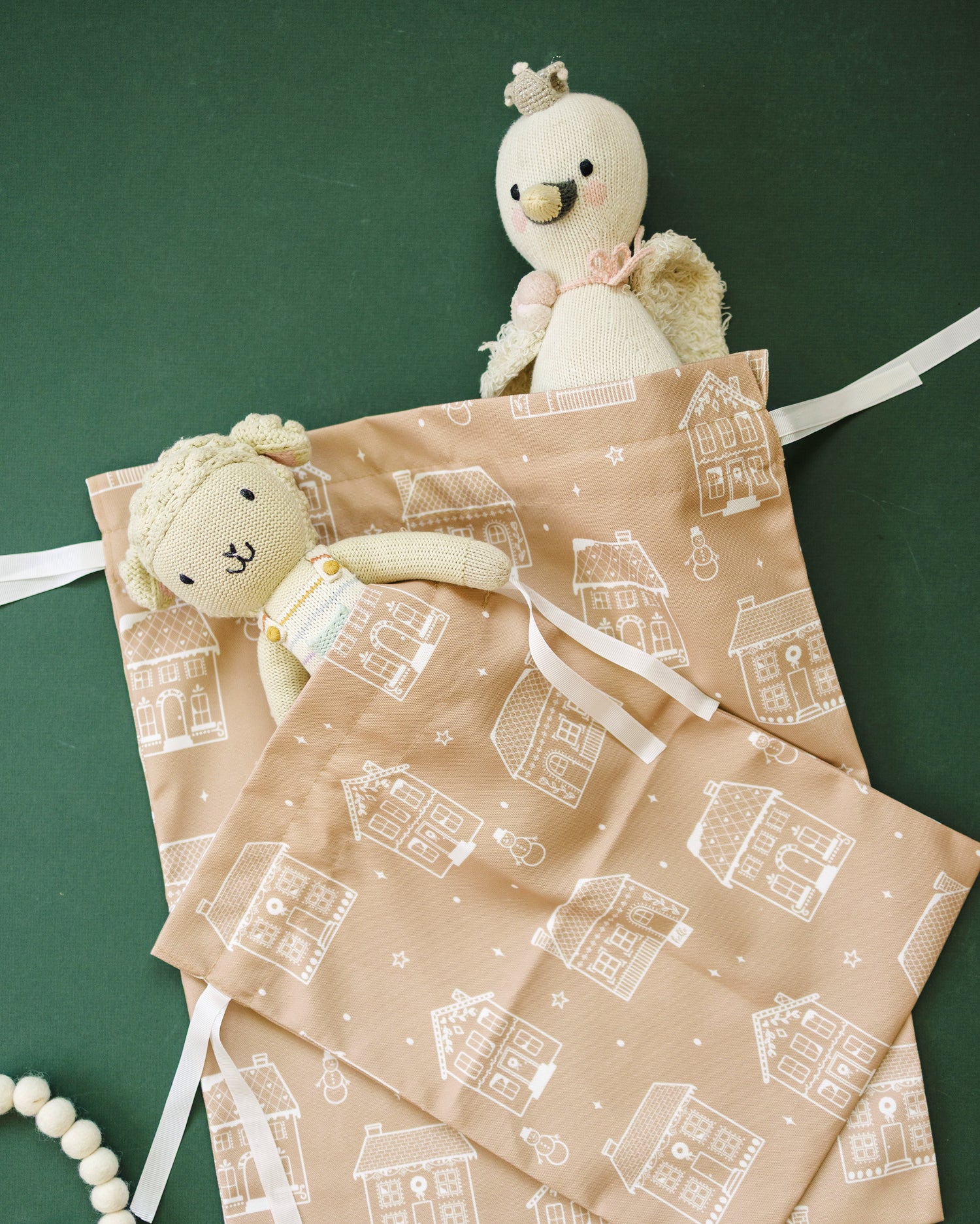 A set of two gingerbread fabric gift bags with a white house pattern, styled with plush toys on a dark green background.