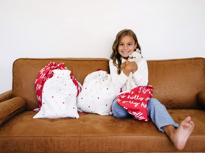 A young girl sits on a brown leather couch, hugging a stuffed toy while sitting by a white patterned holiday fabric gift bag. Two matching fabric storage bags are placed on the couch beside her against a plain white wall.