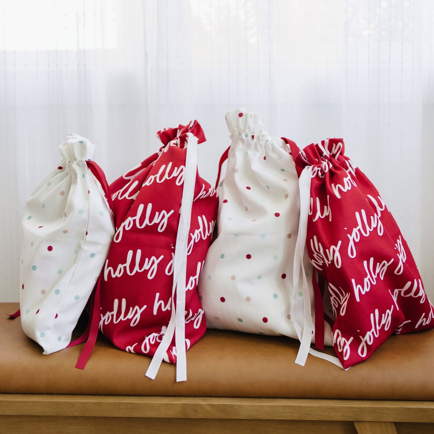 Four fabric drawstring gift bags arranged on a tan bench in front of a white curtain. The bags are white and red with holiday-themed prints, including polka dots and the words 'holly jolly'.