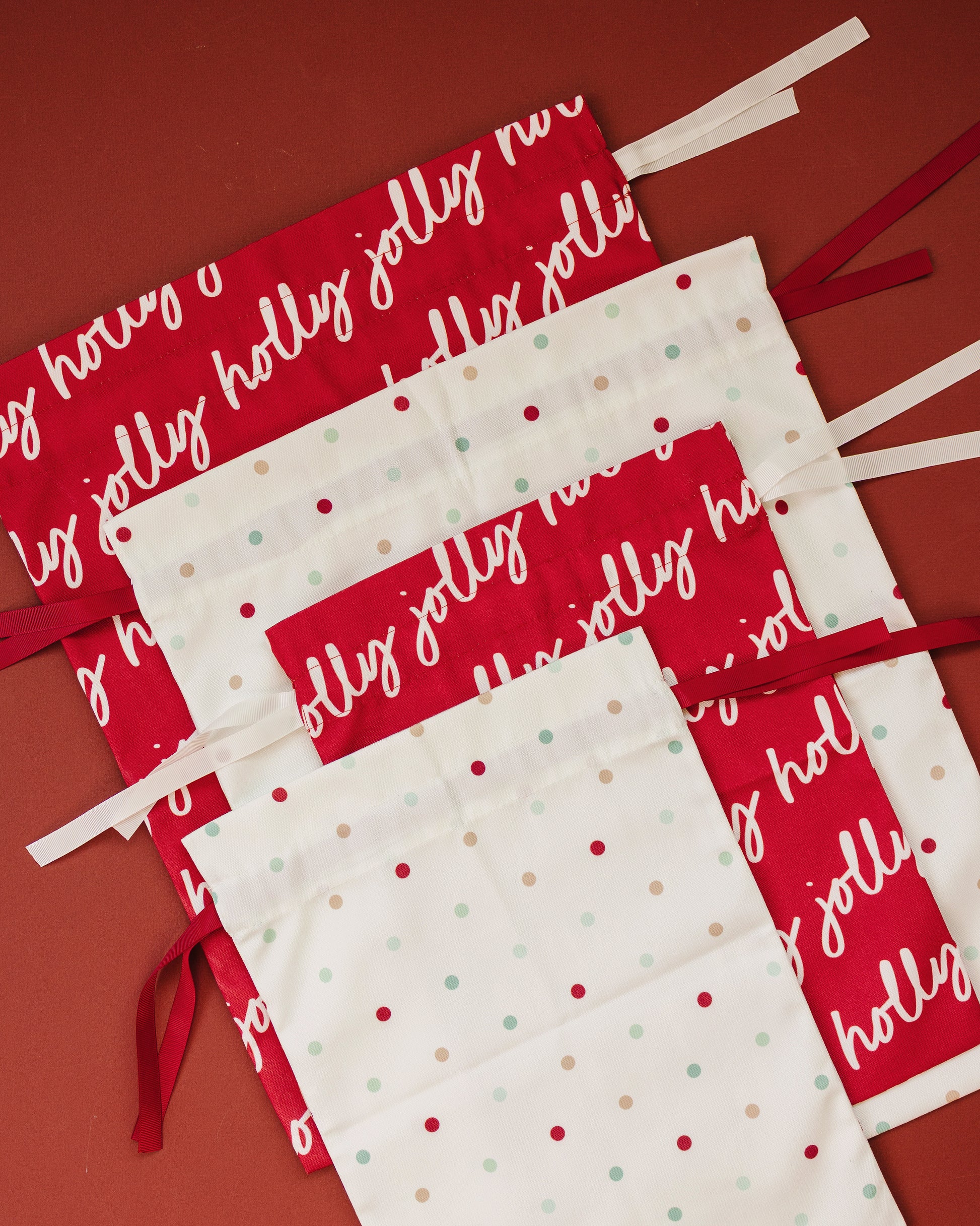 Four drawstring bags arranged diagonally on a solid red background. Two bags are red with white cursive 'holly jolly' text, the other two bags are white with red and mint green polka dots.