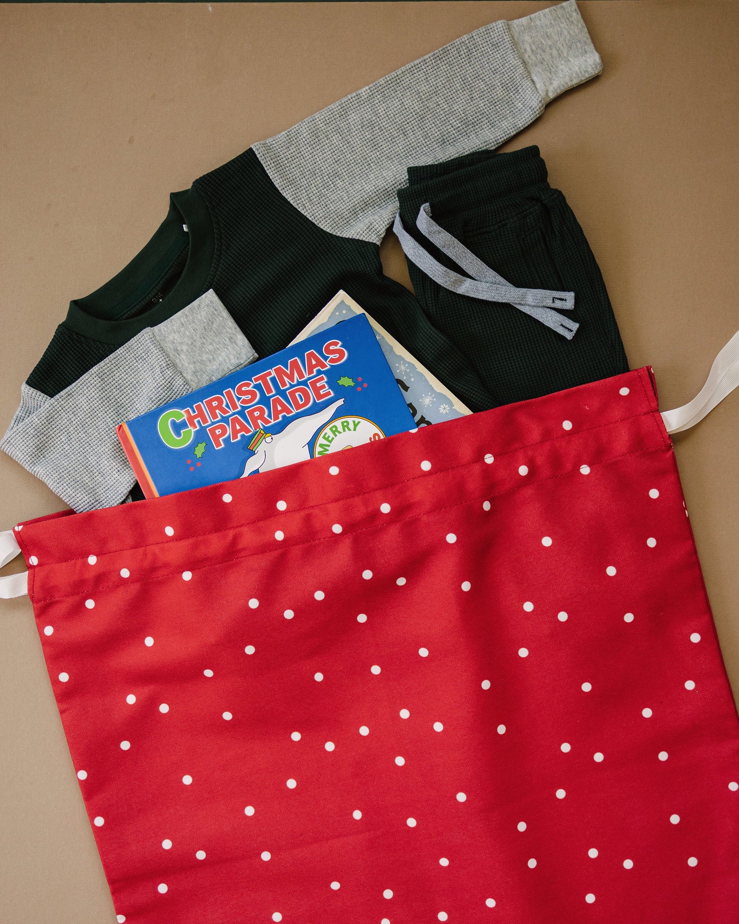 A red drawstring bag with white polka dots filled with folded clothing and a children's book, photographed on a tan background.