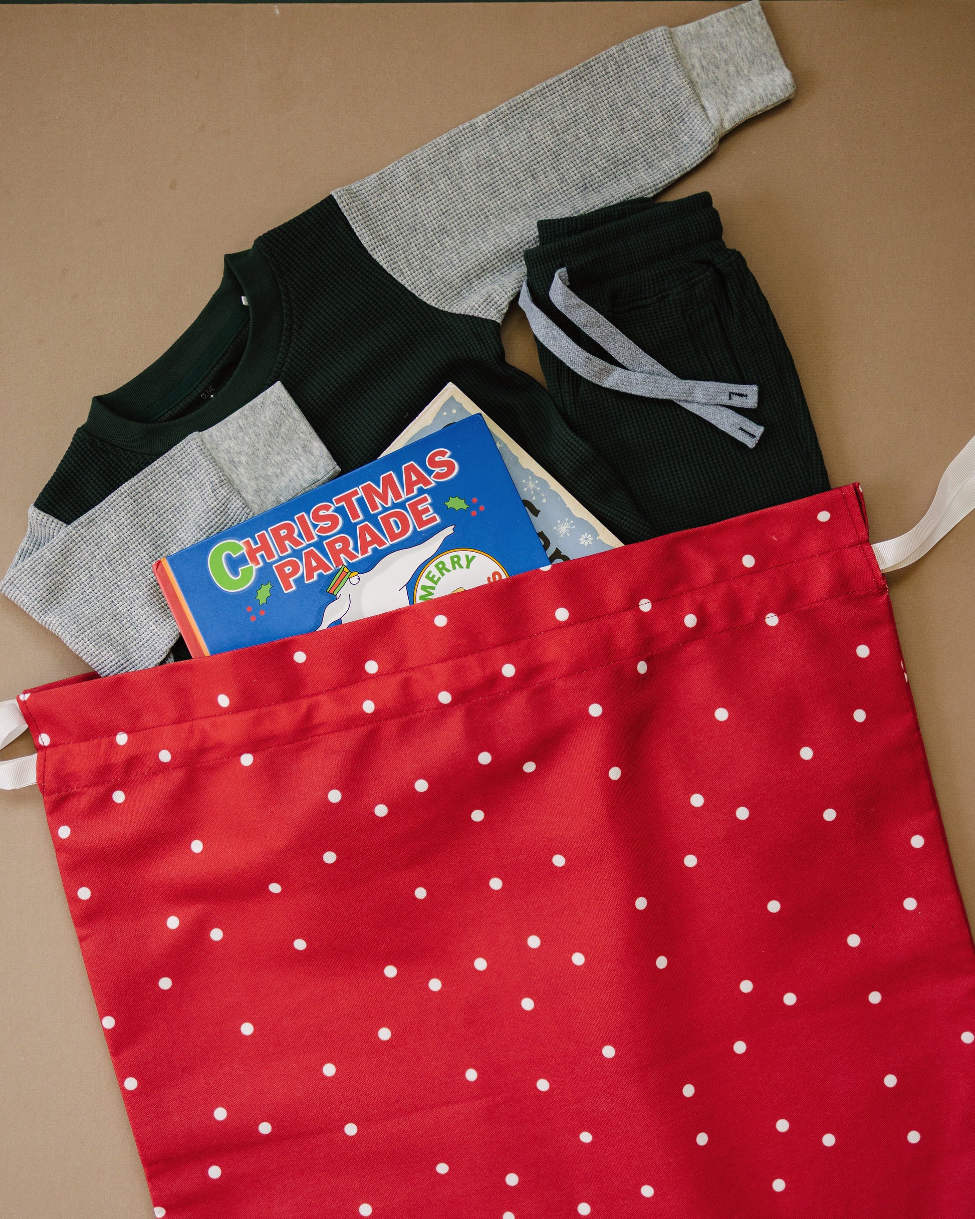 A red drawstring bag with white polka dots filled with folded clothing and a children's book, photographed on a tan background.