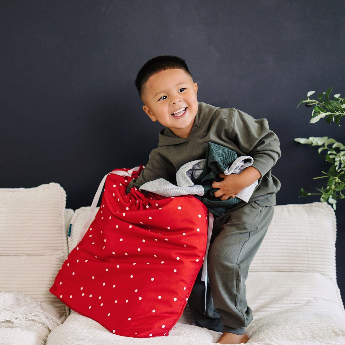 A young boy smiling while sitting on a white sofa, holding a large red laundry bag with white polka dots and a stack of folded clothes against a dark gray wall.