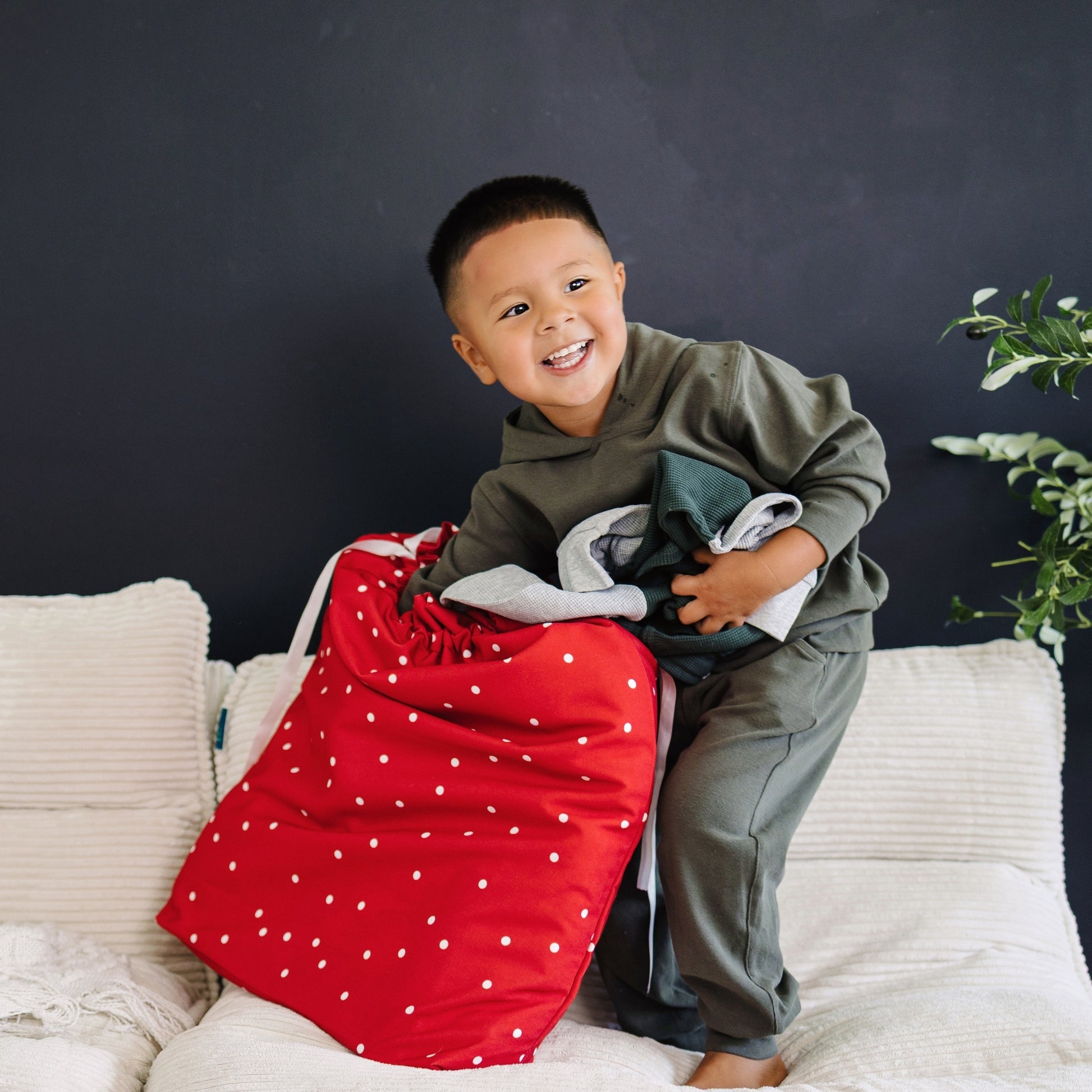 A young boy smiling while sitting on a white sofa, holding a large red laundry bag with white polka dots and a stack of folded clothes against a dark gray wall.