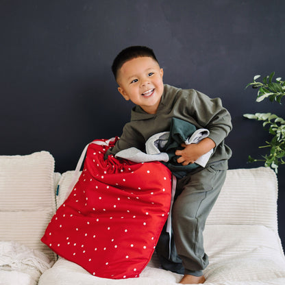 A young boy smiling while sitting on a white sofa, holding a large red laundry bag with white polka dots and a stack of folded clothes against a dark gray wall.