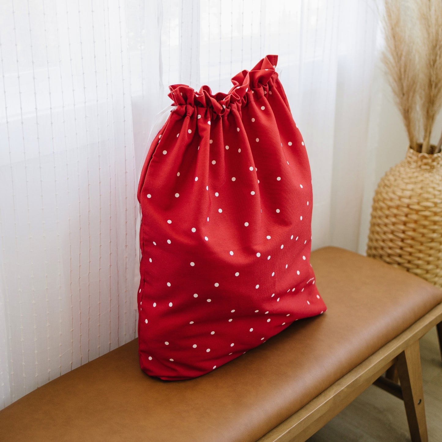 A red drawstring bag with white polka dots sits on a brown leather bench in a softly lit room with sheer curtains and a woven vase in the background.