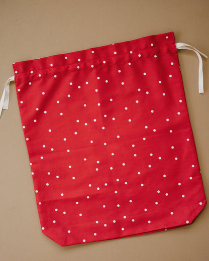 A red fabric drawstring gift bag with a white polka dot pattern, laid flat on a solid tan background.