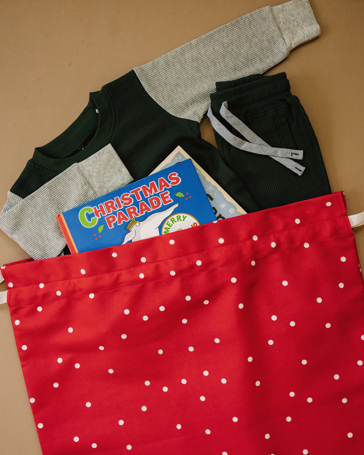 A red fabric gift bag with white polka dots filled with holiday-themed items, including a gray and green long-sleeve shirt, dark pants, a book titled 'Christmas Parade,' and a small gift box, arranged on a tan background.